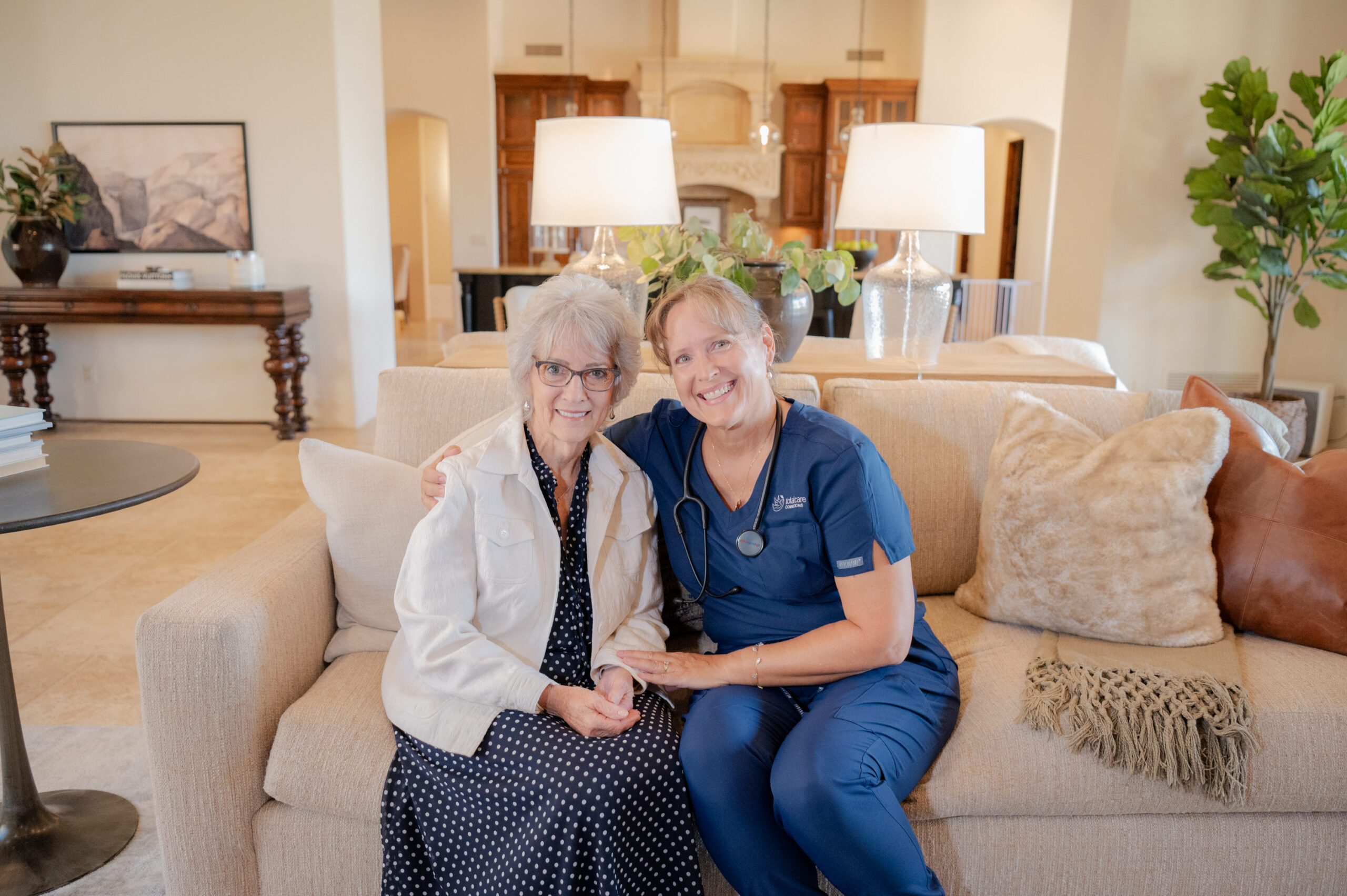 A caregiver from Total Care Connections and a senior client sitting on a sofa, smiling at the camera.